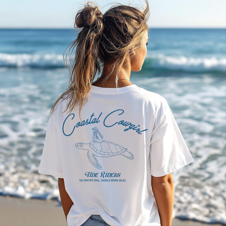 May include: White t-shirt with a blue graphic of a turtle with a person sitting on top of it. The text 'Coastal Cowgirl' is written in blue cursive above the turtle. Below the turtle is the text 'Tide Riders' and 'Saltwater Soul. Saddle Worn Soles'.