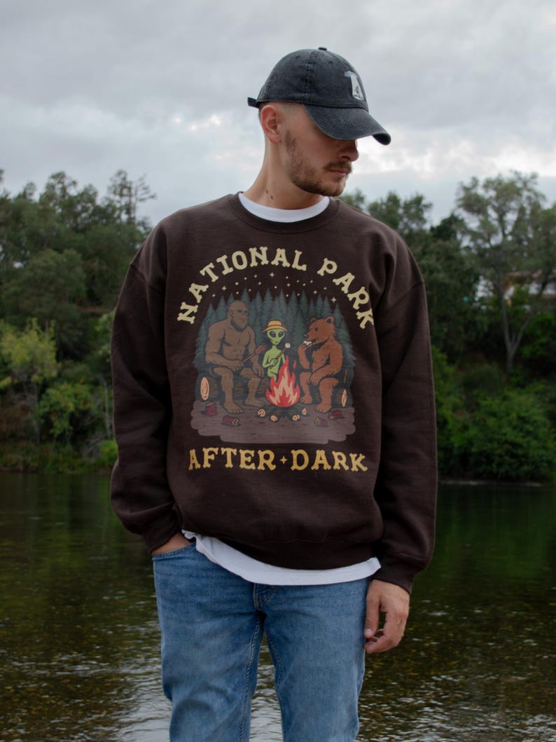 Pode incluir: Uma camisola de gola redonda castanho-escura com uma impress&atilde;o gr&aacute;fica de um Bigfoot, um alien&iacute;gena e um urso sentados &agrave; volta de uma fogueira. O texto "NATIONAL PARK" est&aacute; arqueado acima e "AFTER DARK" impresso abaixo em amarelo.