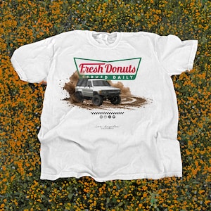 Puede incluir: Camiseta blanca con un gráfico de un vehículo todoterreno plateado levantando polvo, con el texto "Fresh Donuts Served Daily" en un diseño verde y rojo. Debajo, las palabras "Los Angeles, California".
