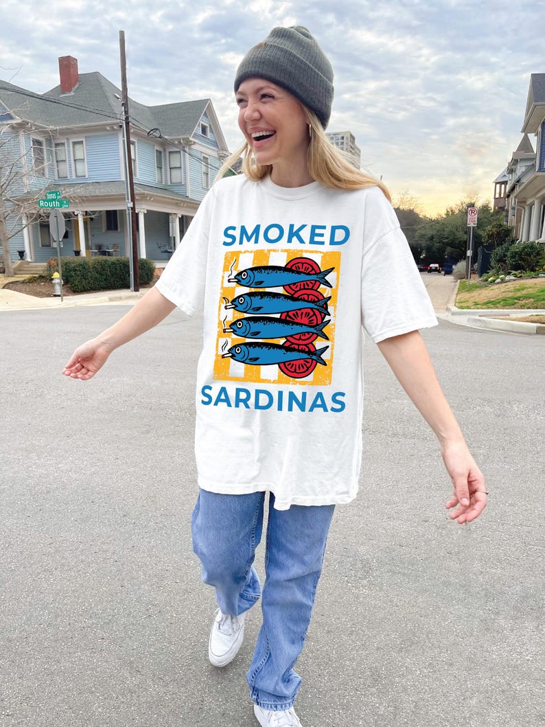 May include: A white t-shirt with a graphic of a stack of smoked sardines on a yellow and white striped background. The text 'SMOKED' is above the sardines and the text 'SARDINAS' is below the sardines.