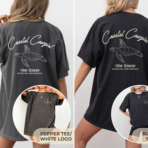 May include: Two oversized t-shirts with a graphic of a sea turtle and the text "Coastal Cowgirl Tide Riders Saltwater Soul Saddle Worn Soles". The left shirt is a pepper color with a white logo and the right shirt is black with a tan logo.