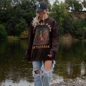 Pode incluir: Uma camisola de gola redonda castanho-escura com um gr&aacute;fico de uma fogueira com um urso, um alien&iacute;gena e um sasquatch, com o texto National Park After Dark. A camisola &eacute; combinada com cal&ccedil;as de ganga azul-claras rasgadas e um bon&eacute; cinzento-escuro.