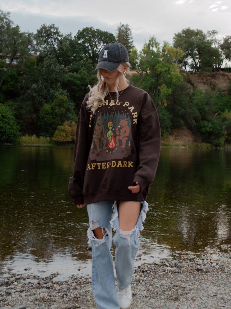 Pode incluir: Uma camisola de gola redonda castanho-escura com um gr&aacute;fico de uma fogueira com um urso, um alien&iacute;gena e um sasquatch, com o texto National Park After Dark. A camisola &eacute; combinada com cal&ccedil;as de ganga azul-claras rasgadas e um bon&eacute; cinzento-escuro.