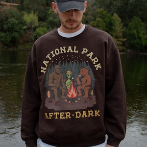 Pode incluir: Uma camisola de gola redonda castanho-escura com um gr&aacute;fico de Bigfoot, um alien&iacute;gena e um urso a assar marshmallows numa fogueira. O texto "NATIONAL PARK AFTER DARK" est&aacute; impresso numa fonte bege acima e abaixo da ilustra&ccedil;&atilde;o.