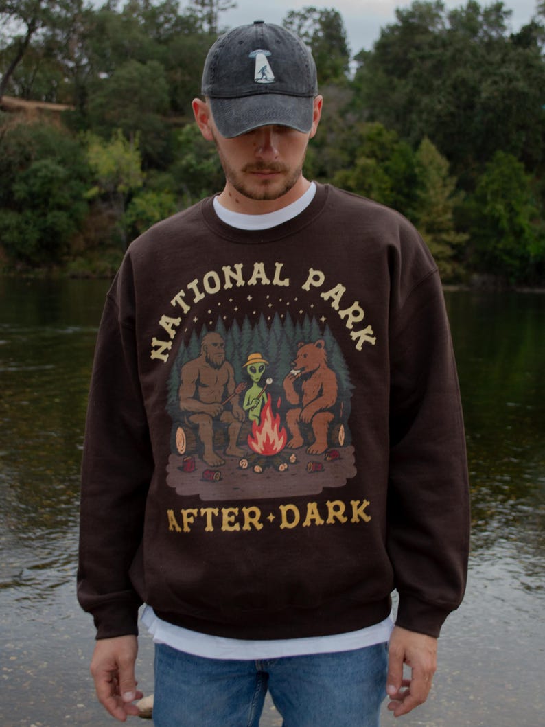 Pode incluir: Uma camisola de gola redonda castanho-escura com um gr&aacute;fico de Bigfoot, um alien&iacute;gena e um urso a assar marshmallows numa fogueira. O texto "NATIONAL PARK AFTER DARK" est&aacute; impresso numa fonte bege acima e abaixo da ilustra&ccedil;&atilde;o.