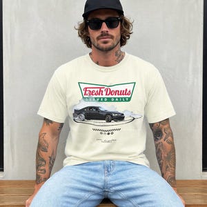 Może przedstawiać: Kremowa koszulka z grafiką czarnego samochodu i napisem "FRESH DONUTS SERVED DAILY" w kolorze zielonym i czerwonym. Koszulka ma również napis "Los Angeles, California" pod grafiką samochodu.