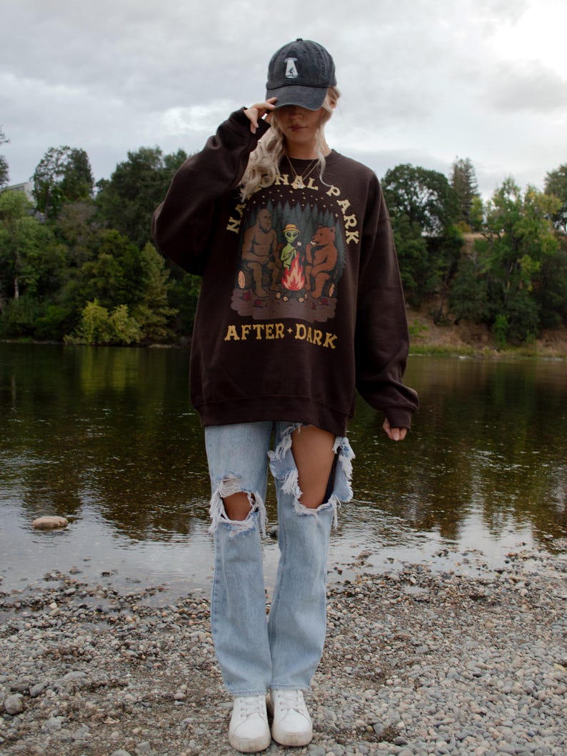 Pode incluir: Uma camisola de gola redonda castanho-escura com um gr&aacute;fico de Bigfoot, um alien&iacute;gena e um urso sentados &agrave; volta de uma fogueira com o texto NATIONAL PARK AFTER DARK, combinada com cal&ccedil;as de ganga azul-claras rasgadas e um bon&eacute; cinzento-escuro.