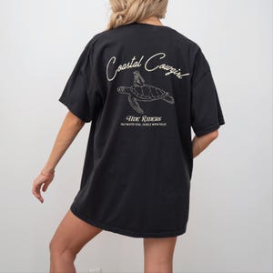 May include: Black oversized t-shirt with a graphic of a sea turtle and the text "Coastal Cowgirl Tide Riders Saltwater Soul, Saddle Worn Soles".
