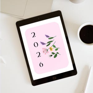 May include: A black tablet showing a pink screen with the numbers "2026" and floral illustrations. The tablet is on a white surface, with a laptop, a cup of coffee, a pen, and a decorative vase in the background.