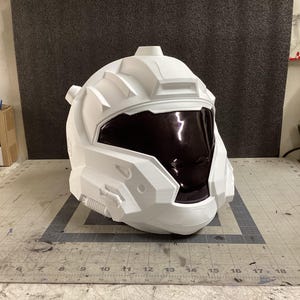 Puede incluir: Un casco blanco de estilo futurista con una visera oscura. El casco tiene un diseño angular complejo con varios paneles y detalles, lo que sugiere un tema de ciencia ficción o cosplay. El casco está sobre una superficie gris.