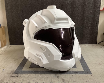 May include: A white, futuristic-style helmet with a dark visor. The helmet has a complex, angular design with various panels and details, suggesting a sci-fi or cosplay theme. The helmet is sitting on a gray surface.