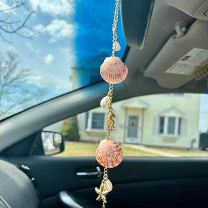 May include: A gold chain with seashells, starfish, and seahorse charms hangs from a rearview mirror in a car. The seashells are pink and white, and the charms are gold.