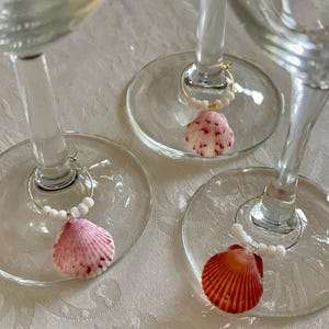 May include: Three wine glasses with seashell charms. Each charm has a small, colourful seashell attached to a metal ring with white beads. The seashells are pink and red, adding a coastal touch to the glassware.