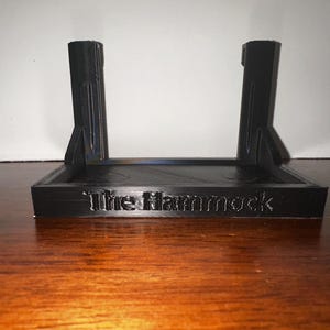 May include: Black 3D-printed stand with the text "The Hammock" on the front. The stand has two vertical supports and a rectangular base. The stand is designed to hold an item. The stand is on a wooden surface.