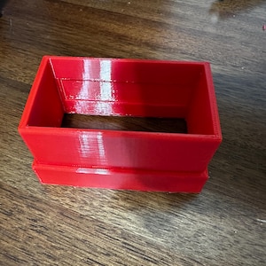 May include: A bright red rectangular container with an open top and bottom. The container is made of a solid material and has a smooth, glossy finish. The object is sitting on a wooden surface.