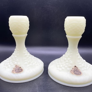 May include: Pair of vintage Fenton candle holders in a creamy, off-white color. Each holder features a textured, bubble-like pattern on the base and stem, with a decorative top. The Fenton logo is visible on each base.