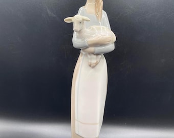 May include: A porcelain figurine of a slender figure with light brown hair, wearing a pale blue top and a long beige skirt, holding a small white lamb in their arms. The figure stands on a smooth, oval-shaped white base with a glossy finish.