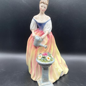 May include: A porcelain figurine of a woman in a long, flowing gown, holding a watering can. The dress is yellow and pink, with white sleeves and a blue bodice. A small pedestal holds a flower arrangement. The figurine is a decorative item.