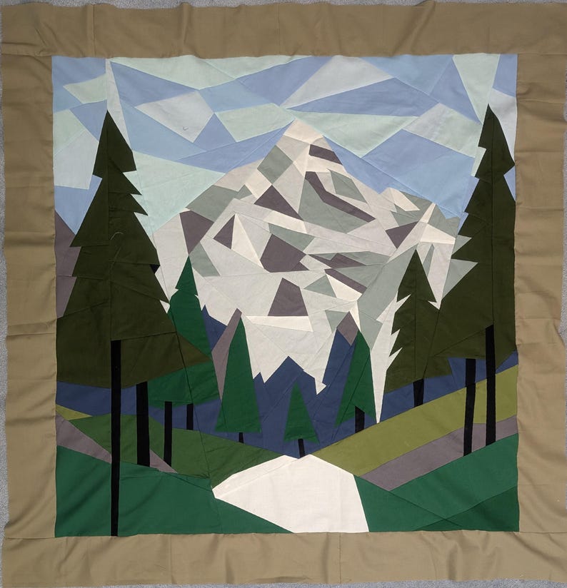 May include: A handmade quilt featuring a mountain landscape design. The quilt showcases a geometric mountain range in shades of gray, white, and blue, with green trees and a light blue sky. The border is a solid tan color.