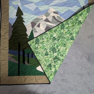 May include: A handmade quilt featuring a mountain and forest scene. The front displays a geometric design of a mountain range, trees, and sky in shades of green, blue, and gray. The quilt's reverse side has a green fern pattern.