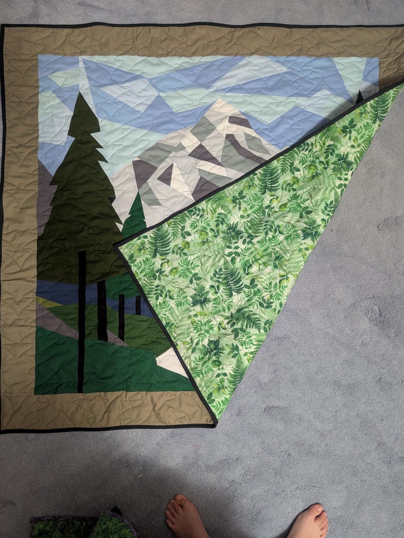May include: A handmade quilt featuring a mountain and forest scene. The front displays a geometric design of a mountain range, trees, and sky in shades of green, blue, and gray. The quilt's reverse side has a green fern pattern.