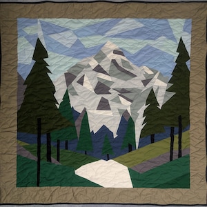 May include: A quilted wall hanging depicting a mountain landscape. The design features a snow-covered mountain, evergreen trees, and a path, in shades of green, blue, grey, and white. It has a brown border and a black trim.