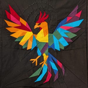 May include: A colorful geometric phoenix design on a black background. The bird features vibrant hues of red, orange, yellow, green, blue, and purple. The wings are spread wide, and the design is reminiscent of a quilt or fabric art.