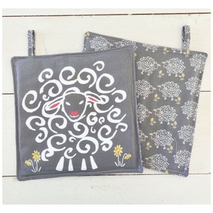 以下が含まれることがあります： 羊のデザインが施されたグレーの綿製鍋つかみ2枚。1枚は、黒い顔と赤い鼻をした白い羊のデザイン。もう1枚は、グレーの背景に黒い顔と赤い鼻をした白い羊が繰り返し描かれたデザインです。