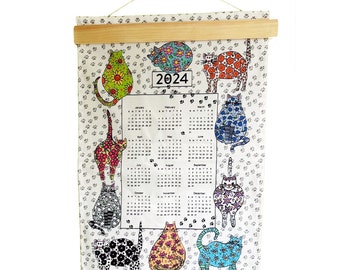 May include: A 2024 calendar featuring cats with colorful floral patterns. The calendar is printed on a white fabric with a gray paw print pattern. The calendar is hanging from a wooden dowel rod.