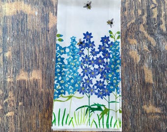 May include: White cotton kitchen towel featuring a floral design with blue and purple flowers and green stems. The towel has a border of green grass and three bees. The towel is folded and placed on a wooden surface.