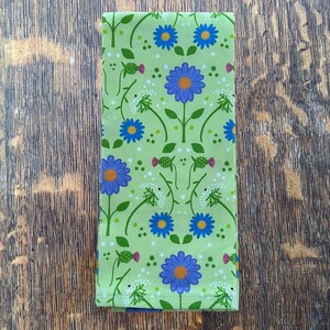 May include: A folded green floral patterned fabric. The fabric features a repeating pattern of blue and orange flowers, white dotted accents, and green stems and leaves. The background is a light green colour.