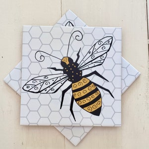 May include: A black and gold bee with intricate details is depicted on a white tile with a honeycomb pattern. The bee has a black body with gold stripes and swirls on its wings.