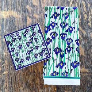 May include: A square coaster and a rectangular towel featuring a floral pattern of purple irises, green stems, and small dragonflies on a white background. The coaster has a dark blue border. The towel has a green and blue stripe at the bottom.