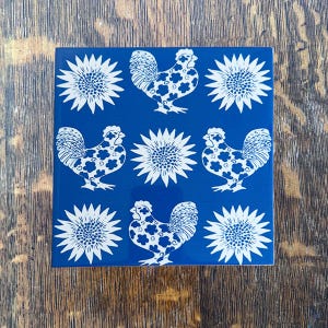 May include: Square blue ceramic tile with a repeating pattern of white sunflowers and roosters. The roosters have a floral pattern on their bodies. The tile is on a wooden surface.
