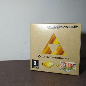 May include: A gold-colored Game Boy Advance SP Zelda Limited Edition Pak box. The front features the Game Boy Advance SP logo, a large gold Triforce symbol, the text Zelda Limited Edition Pak, a 3+ age rating, and a Zelda The Minish Cap logo.