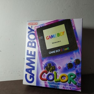 Può includere: Scatola per console di videogiochi Game Boy Color. La scatola bianca presenta la scritta blu "GAME BOY" sul lato sinistro. La parte anteriore mostra una grafica viola della console e le parole "GAME BOY COLOR" a colori.
