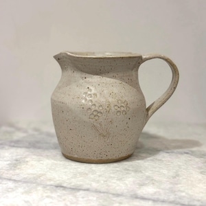 Peut inclure: Un pichet en céramique blanche avec un glaçage moucheté et un motif floral. Le pichet a un corps arrondi et une anse.