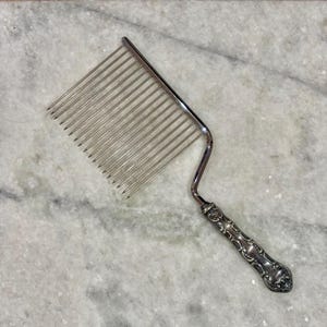 May include: Silver cake slicer with a comb-like blade and ornate handle. The handle features detailed floral and scrollwork designs. The slicer is made of shiny metal and is designed for cutting cakes or pastries. The tool is set against a marble surface.