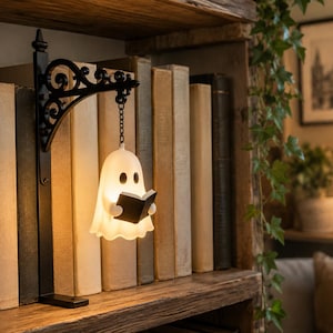 May include: A white, glowing ghost-shaped lamp suspended from a black, ornate metal wall bracket. The ghost figure has two dark oval eyes and holds a small, open black book in its hands. The lamp emits a warm, soft light, creating a gentle illumination.