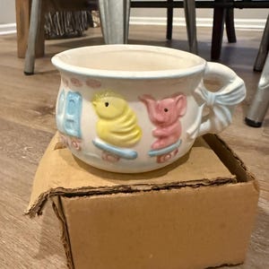 Puede incluir: Una taza de bebé de cerámica blanca con borde y asa azules en forma de lazo. La taza presenta imágenes en relieve de un pollito amarillo y un elefante rosa. La taza está sobre una caja de cartón.