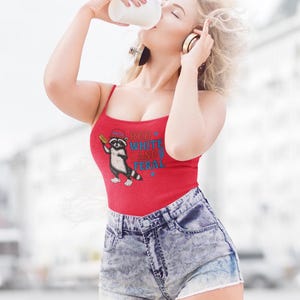 Puede incluir: Una camiseta sin mangas roja con el texto "RED WHITE AND FERAL" y un gráfico de mapache. La modelo lleva pantalones cortos vaqueros y auriculares, sosteniendo una taza blanca. La imagen está tomada al aire libre.