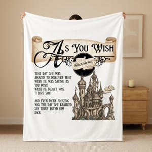 Puede incluir: Manta blanca con el texto "As You Wish" en un diseño de pergamino. Debajo, una ilustración de un castillo y los nombres "Natalie and Nick". La manta incluye una cita romántica.