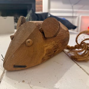 May include: A wooden mouse figurine with a brown rope tail. The mouse has a carved face with a small black nose and two black whiskers.