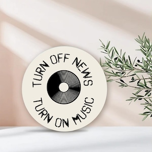 May include: A round, off-white coaster with the text "TURN OFF NEWS TURN ON MUSIC" encircling a black vinyl record graphic. The coaster is set against a neutral background with a sprig of greenery.