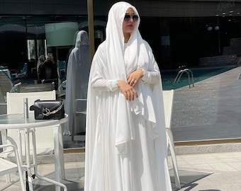 May include: A white abaya with a long, flowing skirt and a matching headscarf. The abaya is made of a soft, silky fabric and has a simple, elegant design. The woman is wearing black sunglasses.