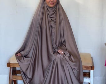 May include: A brown satin abaya with a matching head covering. The abaya is draped over a person seated on a wooden bench. The fabric has a subtle sheen, and the design is modest and flowing.
