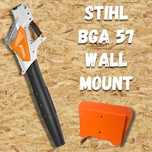 Può includere: Un soffiatore per foglie STIHL BGA 57 bianco e arancione con ugello nero e supporto a parete arancione. Il testo "STIHL BGA 57 WALL MOUNT" è visualizzato in lettere bianche su sfondo effetto legno.