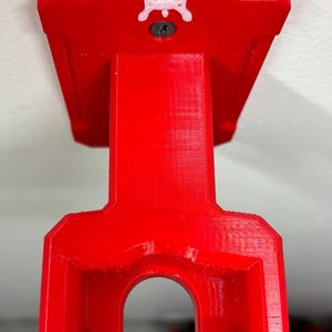 May include: A bright red, 3D-printed wall mount. The mount has a rectangular top with a white emblem and a screw. The bottom has a curved opening. The mount is designed to hold an item.