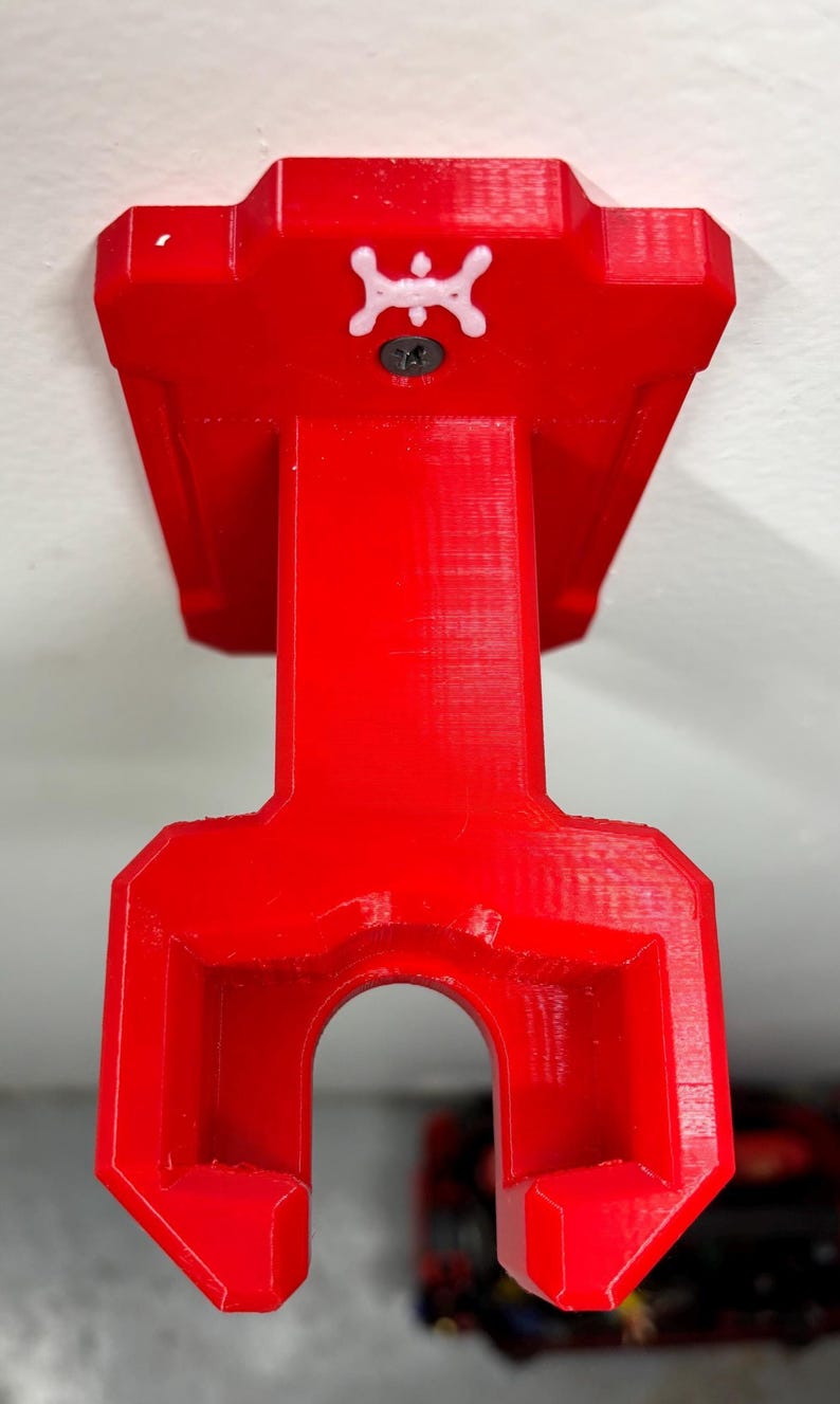 May include: A bright red, 3D-printed wall mount. The mount has a rectangular top with a white emblem and a screw. The bottom has a curved opening. The mount is designed to hold an item.