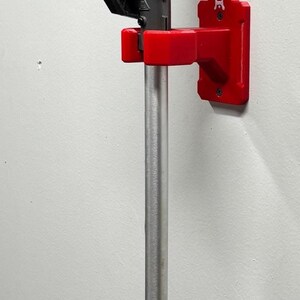 May include: A pole saw mounted on a wall. The saw has a black blade, a tan engine housing, and a silver pole with a black handle. A red bracket holds the saw to the wall.